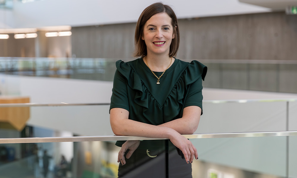 A woman in a green dress leans against a glass railing in Allard Hall.