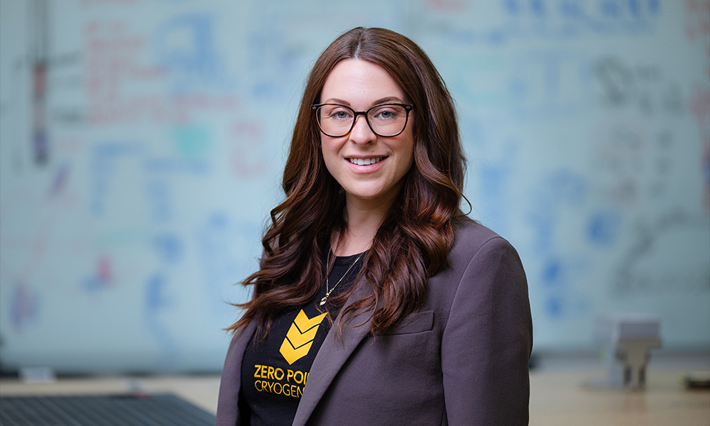 A woman in a blazer smiles, a whiteboard with writing and diagrams blurred behind her.