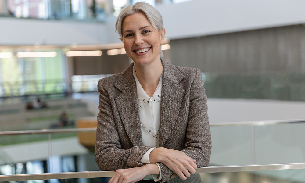 A woman in a brown blazer leans against a glass railing in Allard Hall.