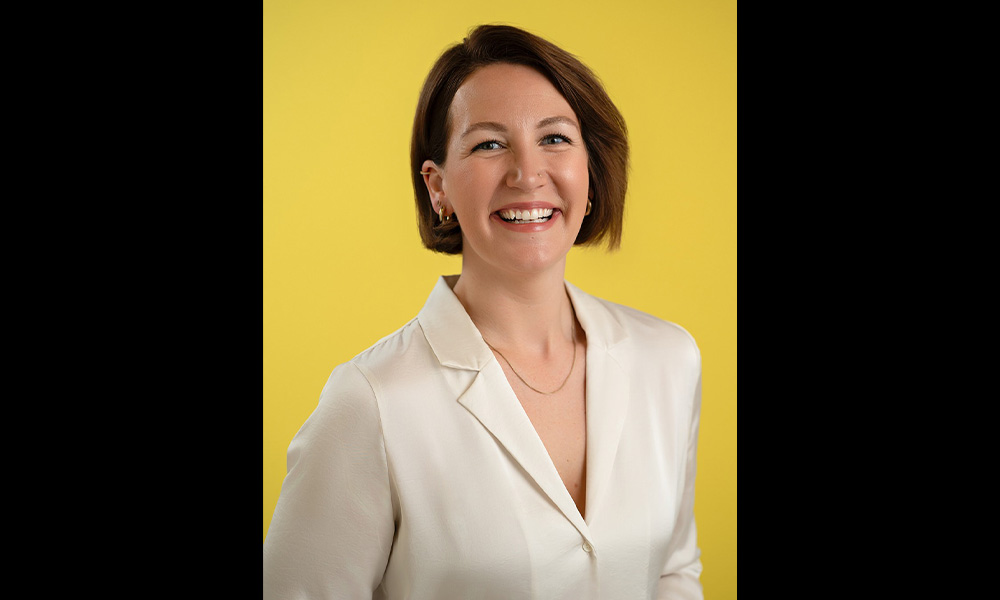 A woman in a white shirt smiles, a bright yellow background behind her.