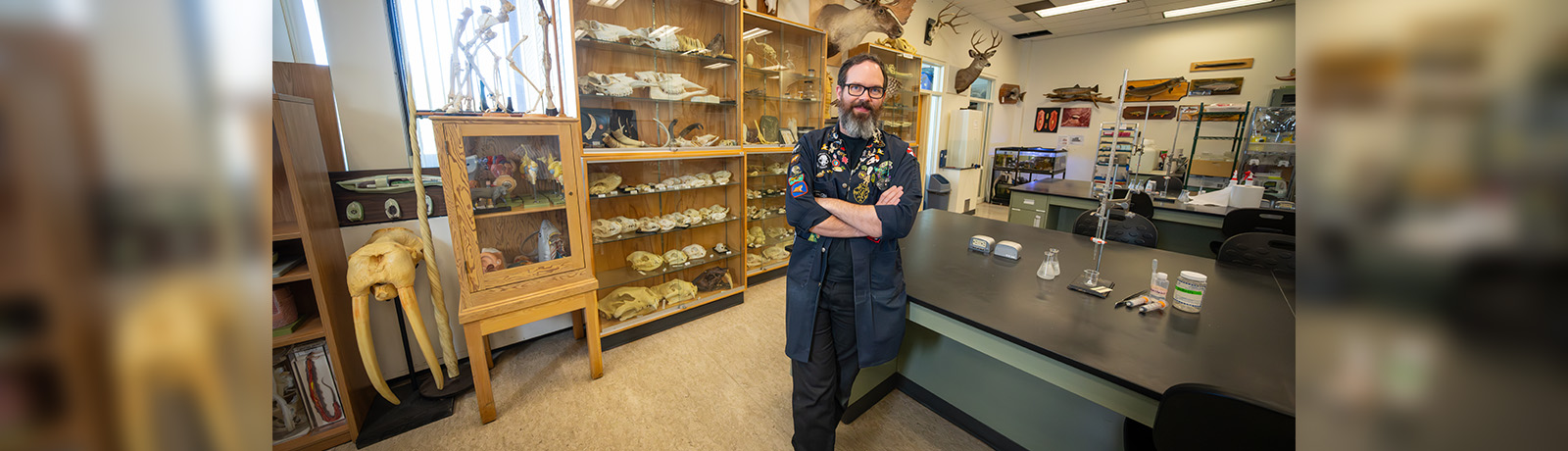 Dana Sanderson leans back against a table in the bio lab, specimens and skeletons on shelves and the wall behind him.