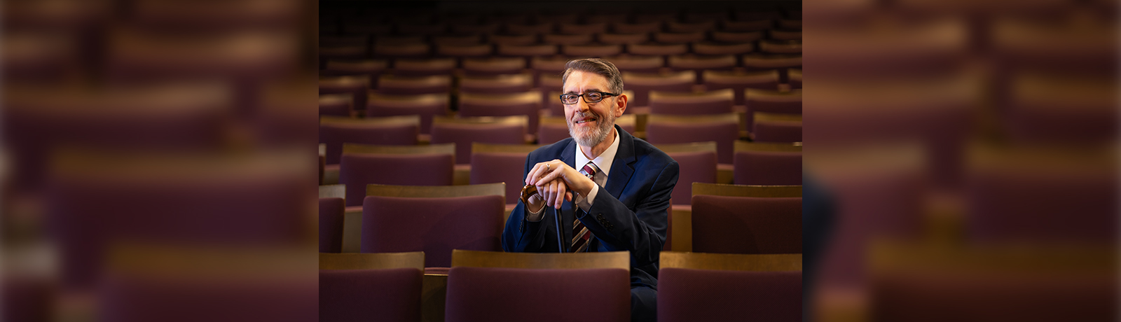 A man sits in a brown theatre seat within an empty theatre