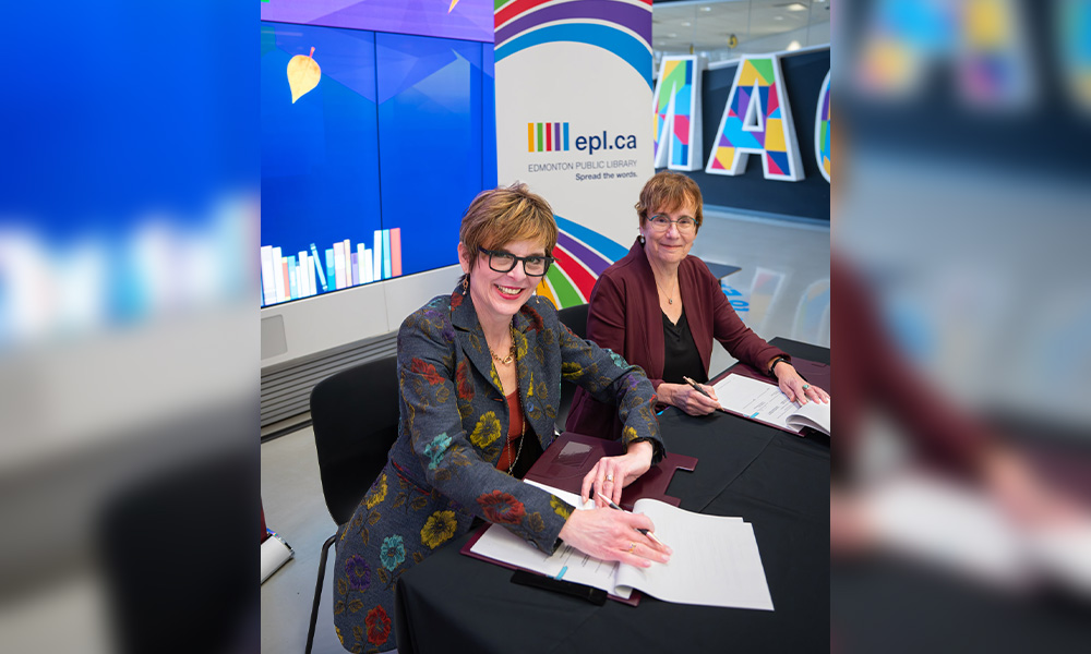 Two women sit at a table, pens in hand to sign a document together.