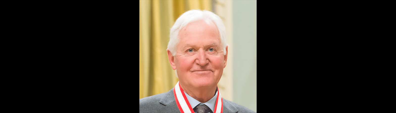 A headshot of a man in a grey suit, with a red and white neckband of a medal visible around his neck.