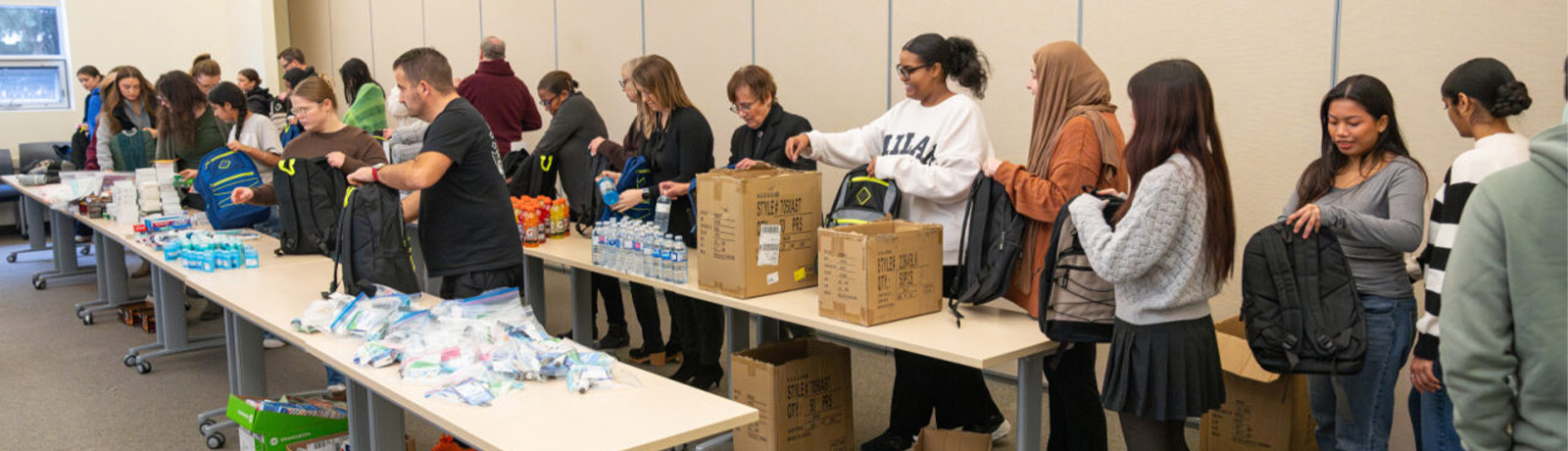 A lineup of students and faculty fill backpacks from stacks of items on classroom tables.