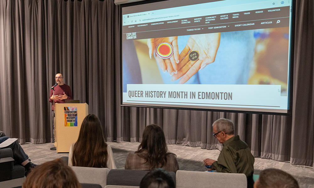 A man stands at a podium, with a large screen next to him reading, "Queer History Month Edmonton."