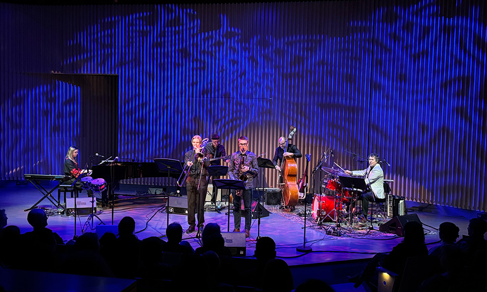 A band plays on a stage with a background of blue interconnecting lines behind them.