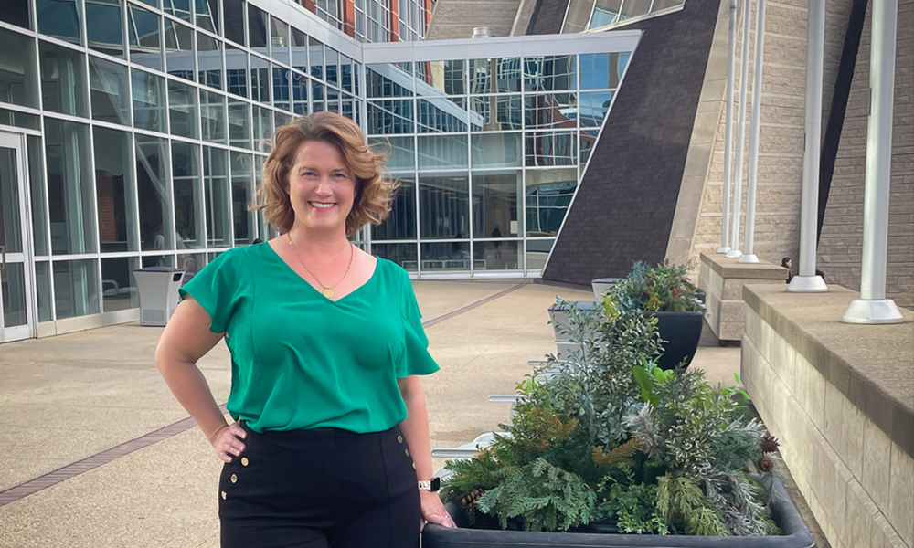 A woman in a green shirt stands in front of glass windows outside of Building 6.