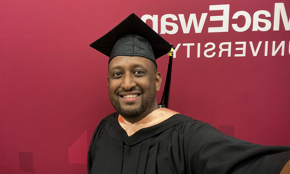 A man wears a black cap and gown in front of a maroon background.