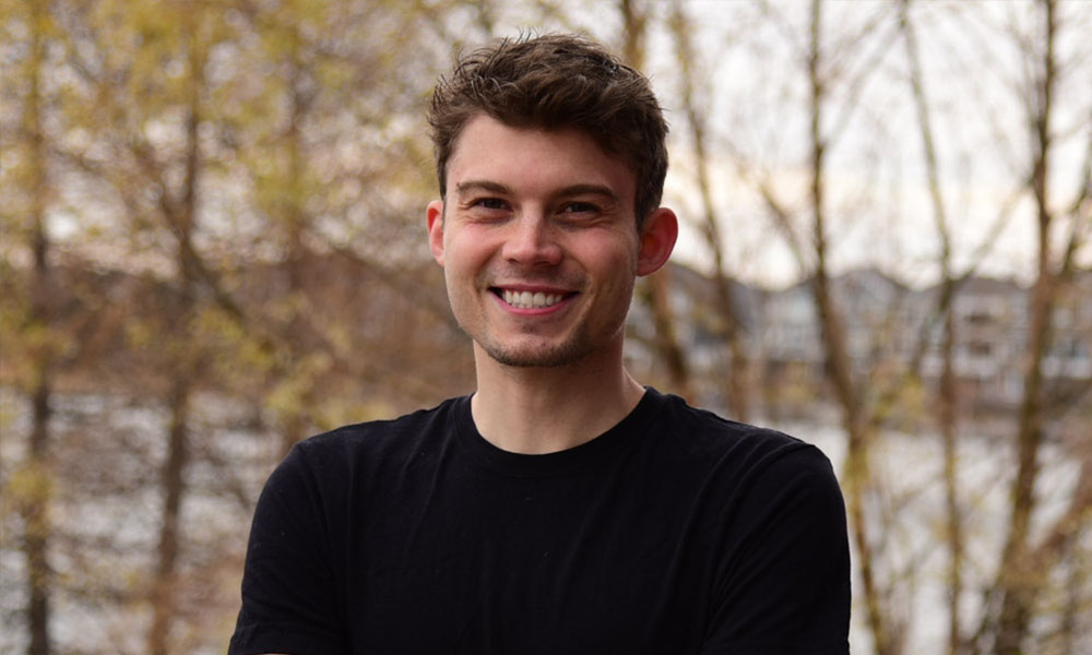 A man in a black shirt smiles, autumn trees blurred behind him.