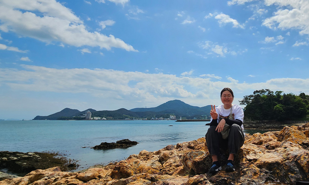 A woman sits on a rocky shore with the ocean behind her, and she gives the peace sign with one hand.