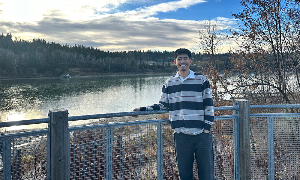 A man stands in front of a wire and wood fence with a river and river bank in the distance