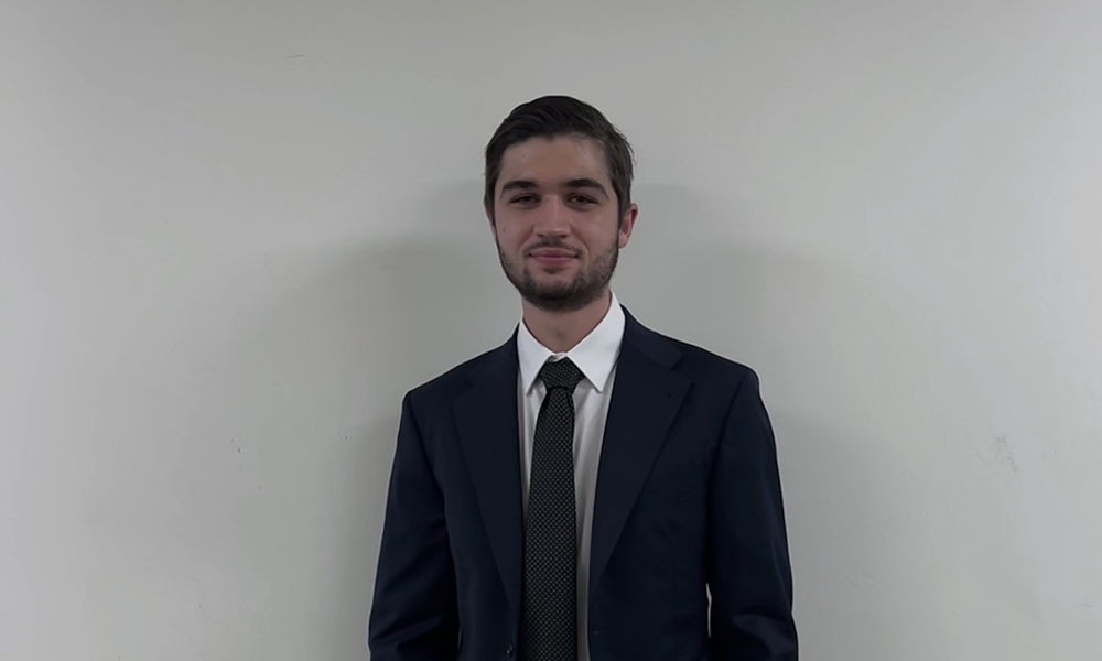 A man in a black suit and tie stands against a white wall.