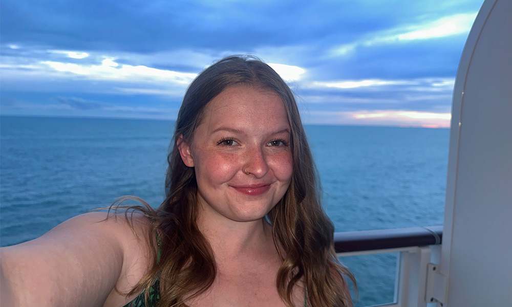 A woman stands against the railing on a boat, the ocean and sunset behind her.
