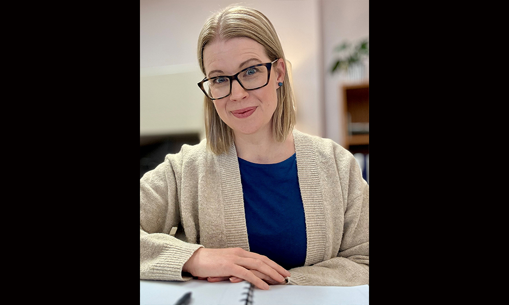 A woman wears a beige cardigan and sits with her hands folded on an open notebook.