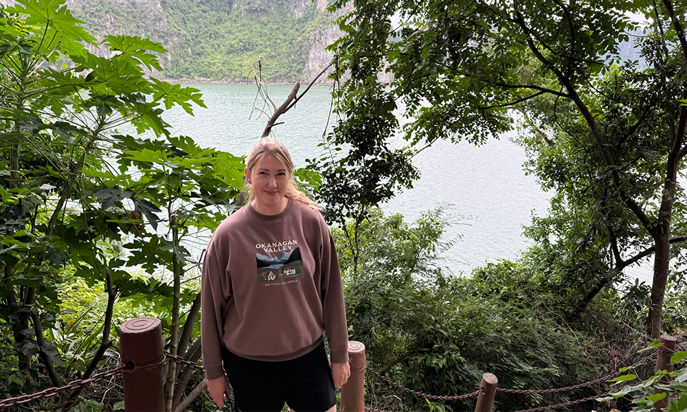 A woman stands in front of a railing on a pathway that is is front of green foliage. There is a body of water behind her.