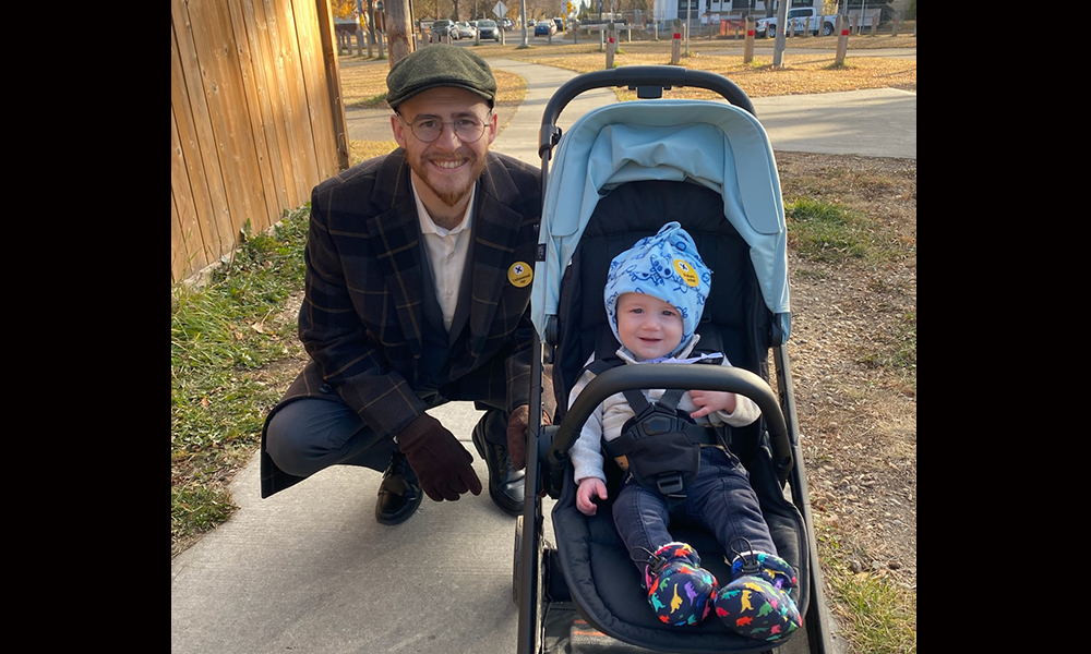 A man in a hat and jacket crouches down on a sidewalk next to a baby in a baby stroller 
