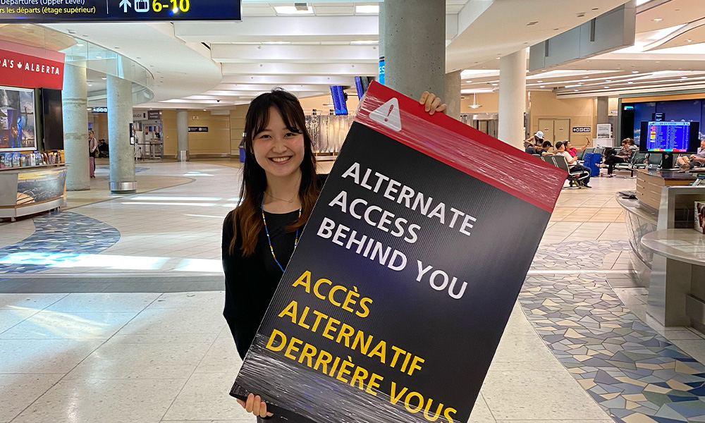 A woman stands in a large corridor, holding a sign that reads alternate access behind you