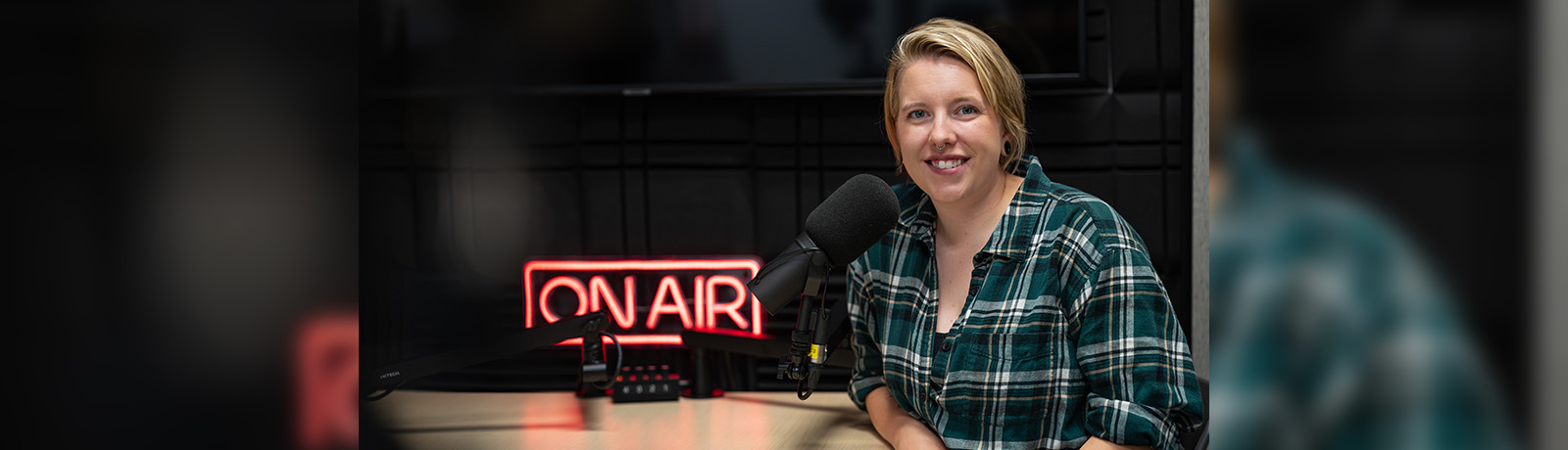 A young woman in a green plaid shirt sits at a table with podcasting equipment and a red "On Air" neon sign illuminated.