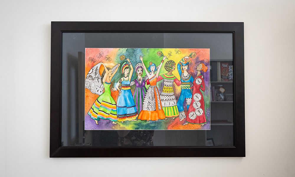 photograph of a framed painting of a group of seven women, all dressed in colourful dresses and headpieces and dancing
