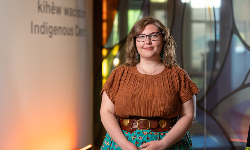 Alix stands with her hands folded in front of her, wearing a brown shirt and a blue skirt.