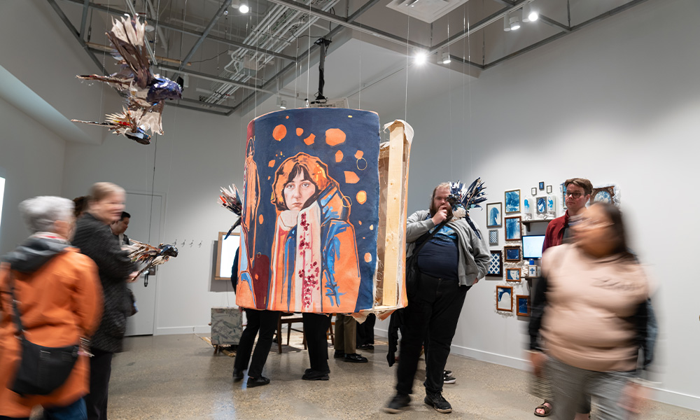 A piece of art hangs in the middle of a room while blurred images of people surround it.