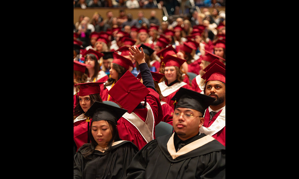 Grads sit together in robes, with one student facing the other way and a hand raised in a wave.