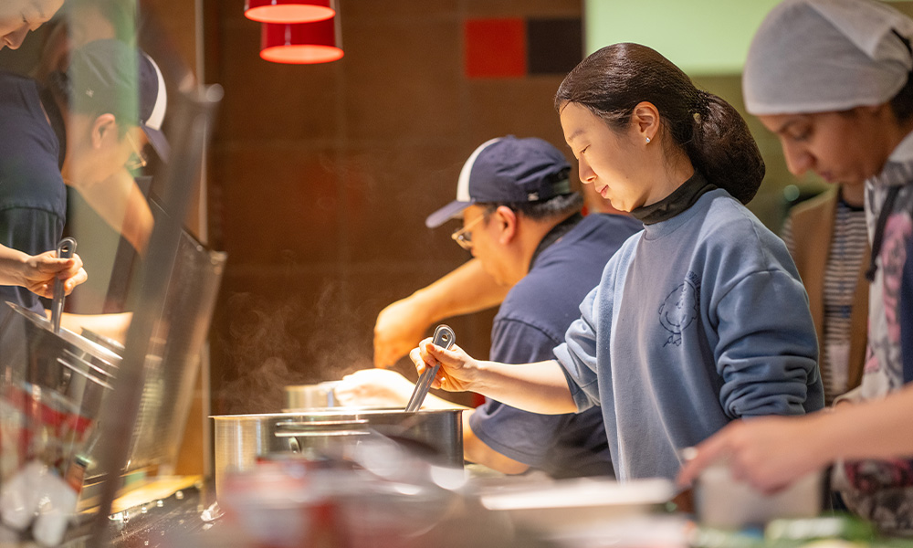 A student stirs something in a pot while two chefs prep food on either side of her.