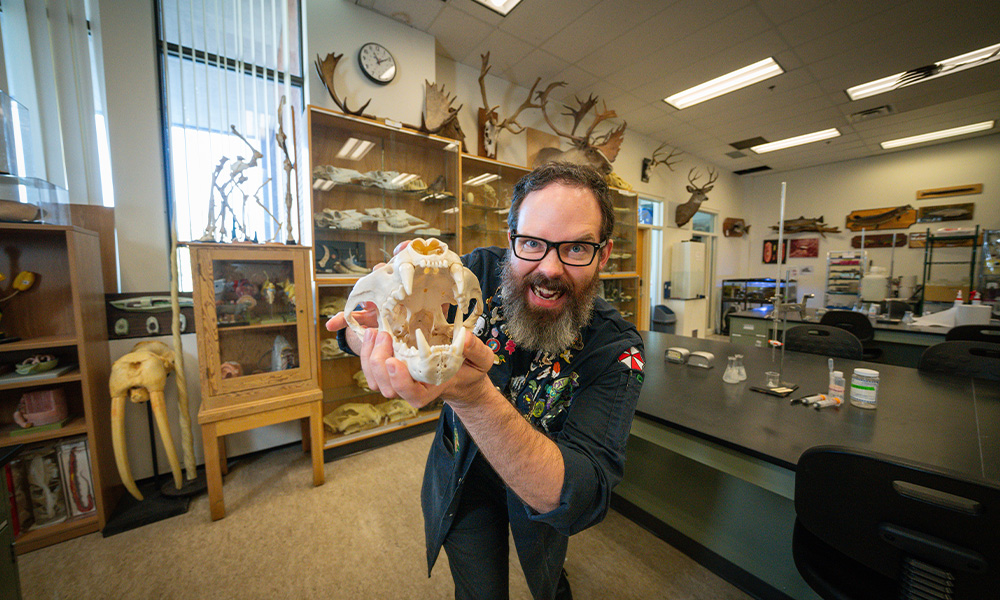 Dana Sanderson stands in the bio lab, holding a cougar skull, baring his teeth at the camera.