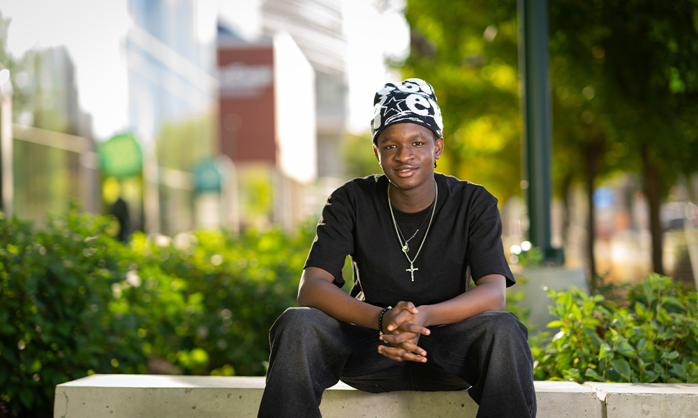 Emmanuel sits on a stone bench outside of Allard Hall with campus blurred behind him.