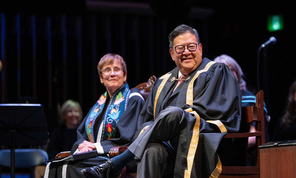 Gary Mar and Dr. Trimbee sit together wearing convocation regalia, smiling.