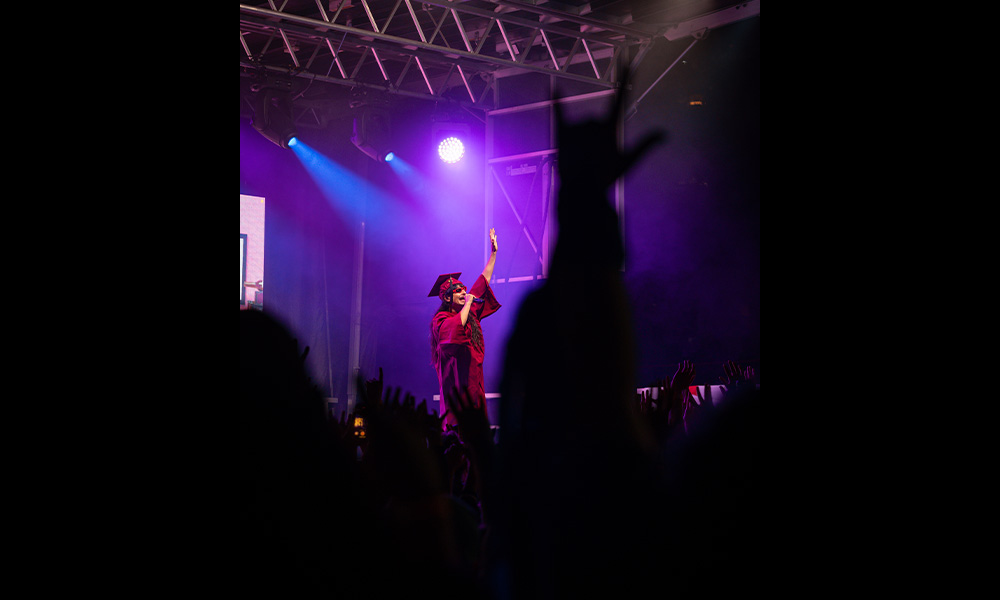 A musician wearing grad robes is on stage with purple lighting, seen from behind raised hands in the crowd.