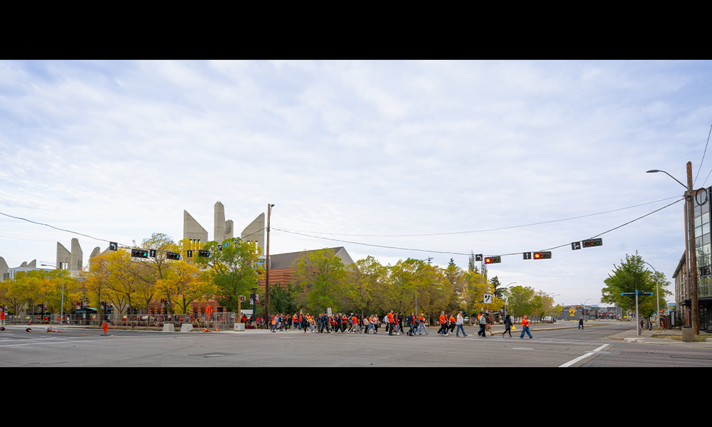 A long line of people are seen crossing the street at an intersection, with MacEwan's towers visible in the far background.