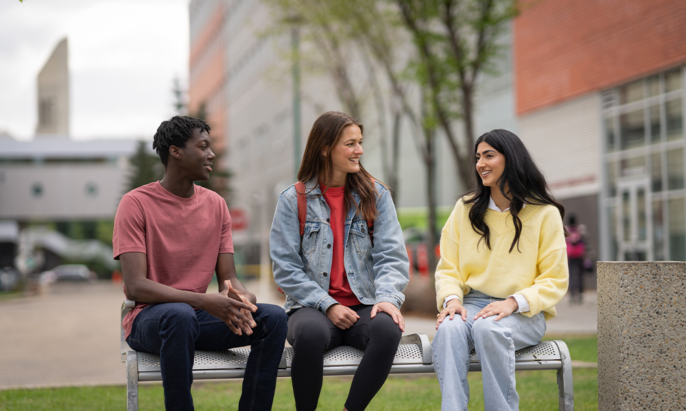 Three students sit on a bench together and chat.
