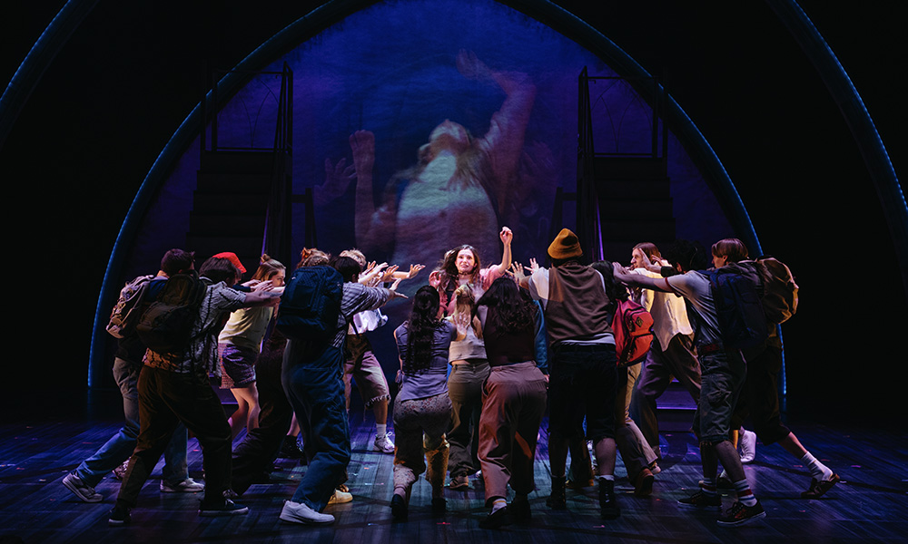A groups of actover crowds around a women with her hand raised against a dark backdrop