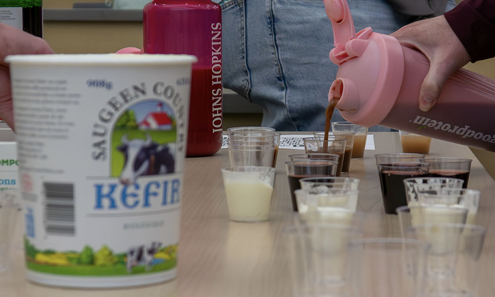 A container of kefir in the foreground against a hand pouring liquids into small cups