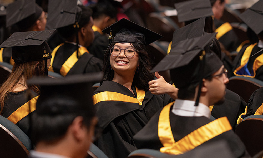 A grad wearing a black gown and sash smiles and gives a thumbs up