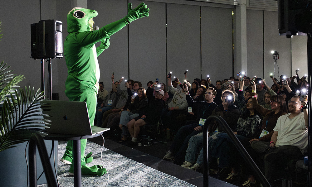 A person dressed in a gecko costume reaches out toward a crowd holding up lit cel phone
