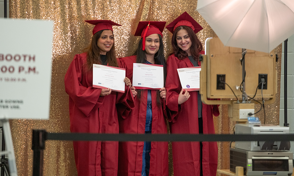 Three people, dressed in red gowns and caps, stand in front of a digital camera holding certificates. 