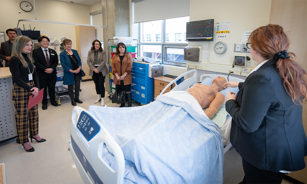 A group of people stand around a hospital bed with a training mannequin in it.