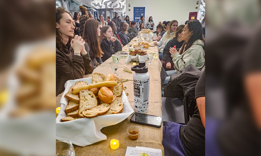 Several students sit at a long table, baskets of bread along the tabletop.