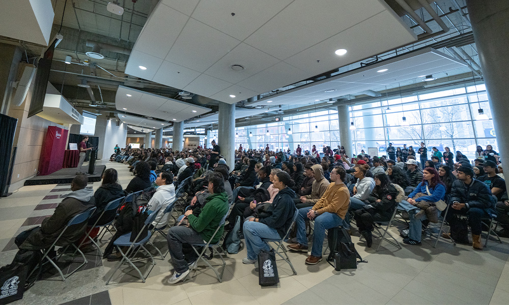 A large crows of new students sit in the Robbins cafeteria, looking toward a speaker at a podium.
