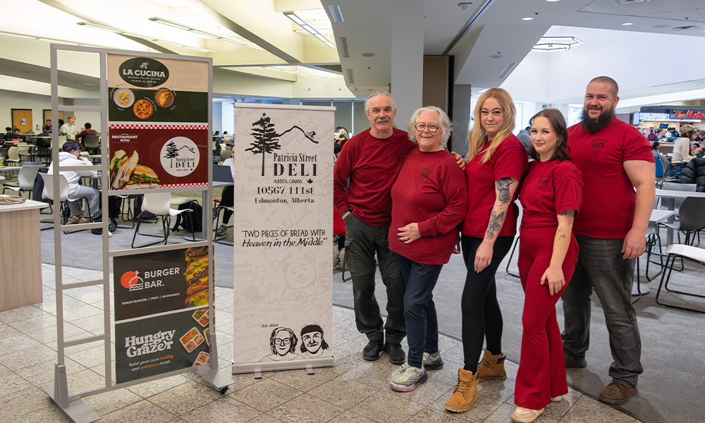 A group of people in red tee shirts stand beside a sign for the Patricia Street Deli in the Building 6 cafeteria.