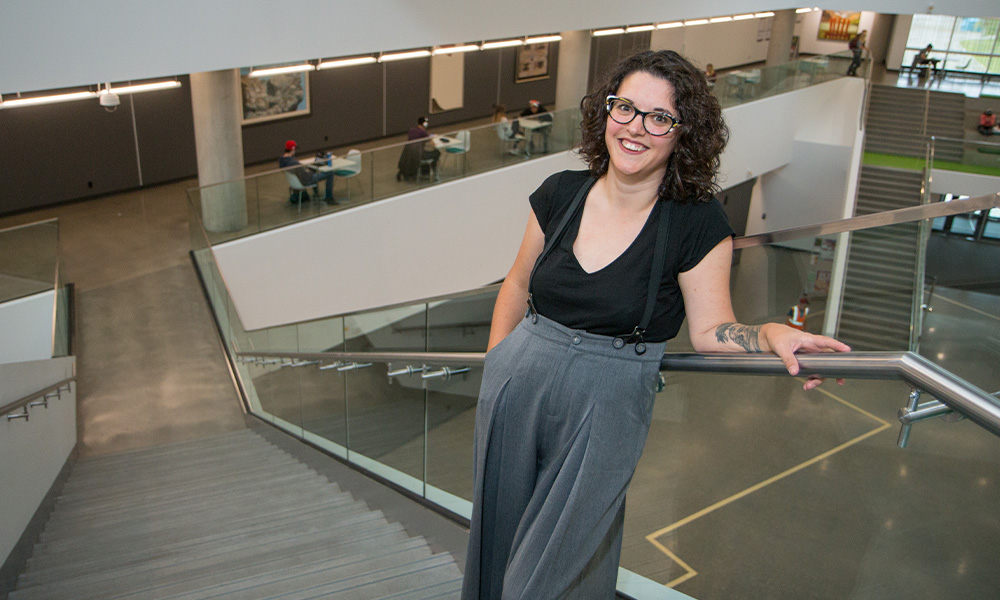 Katie Biittner wears a black tee shirt and patterned pants, and leans back against a railing.
