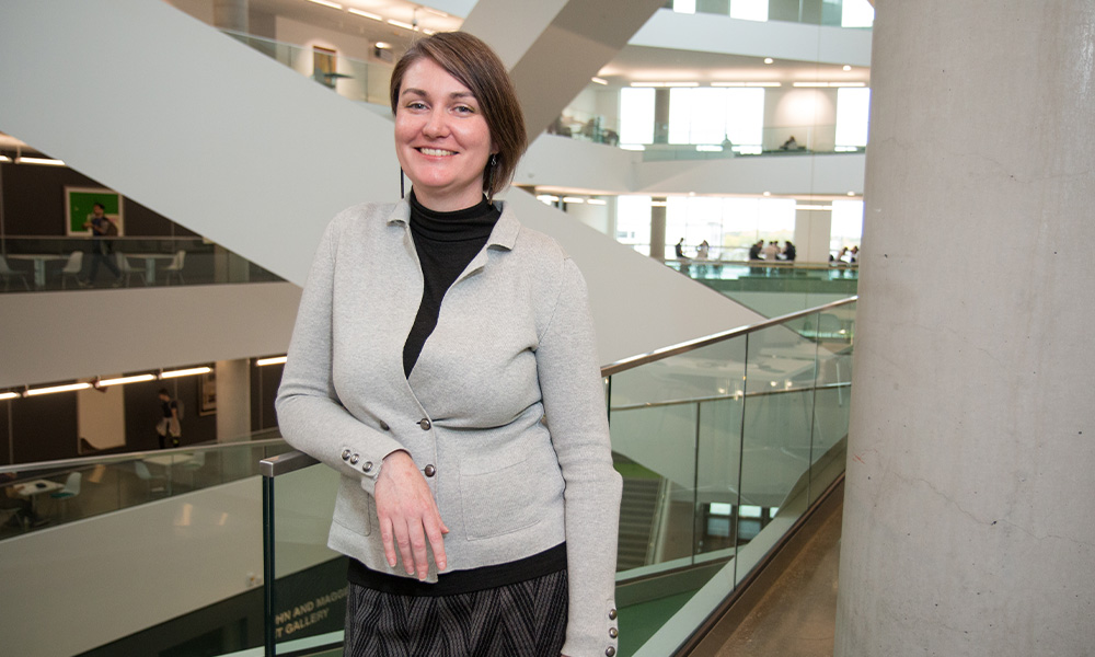 Erin Cowling wears a grey zip-up sweater and leans one arm on a railing.