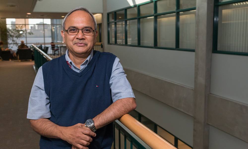 Murli Muralidharan wears a light blue shirt under a dark blue sweater vest, and leans against a railing in Building 5.