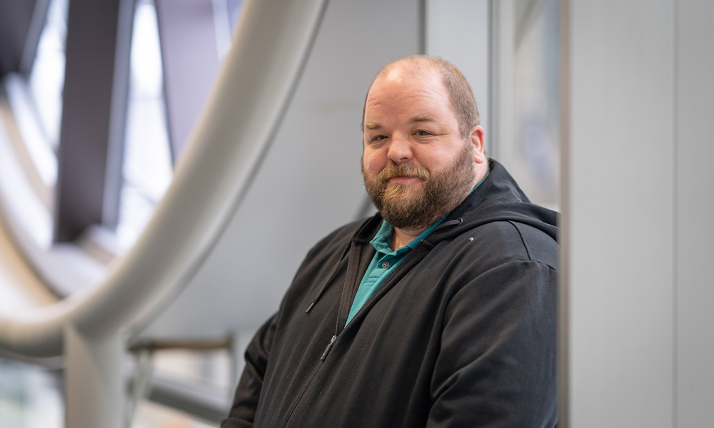 Andrew Seeley wears a grey hoodie and leans against a pillar inside the clocktower window in the library.