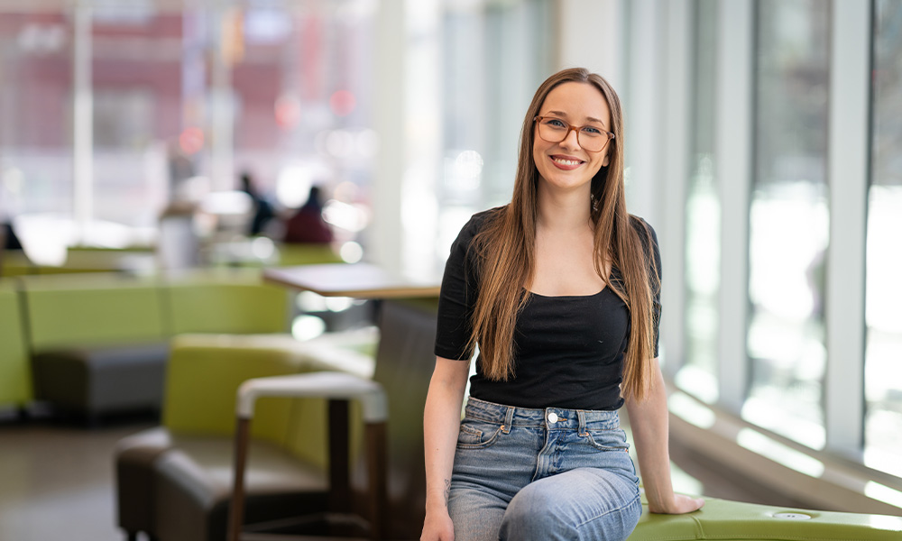 Eva Hollas sits on a green chair in Allard Hall, wearing jeans and a black shirt, smiling.