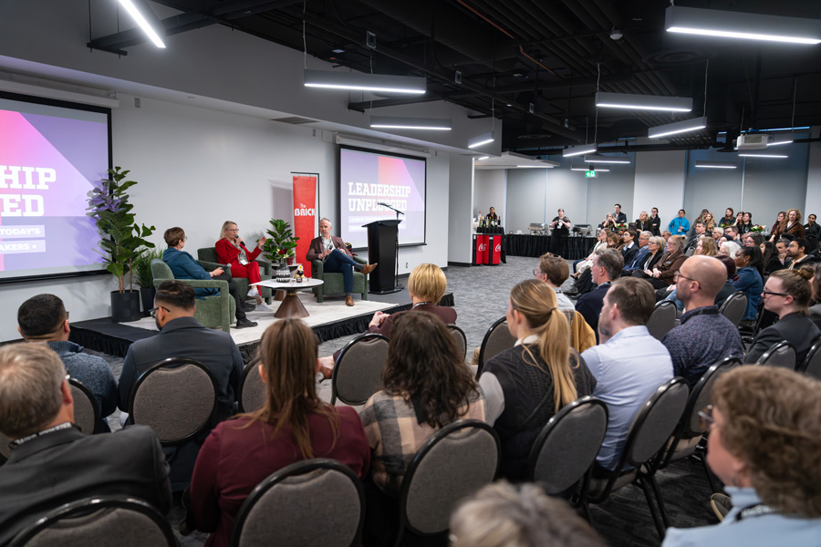 Three speakers sit on a stage at the front of a room filled with people