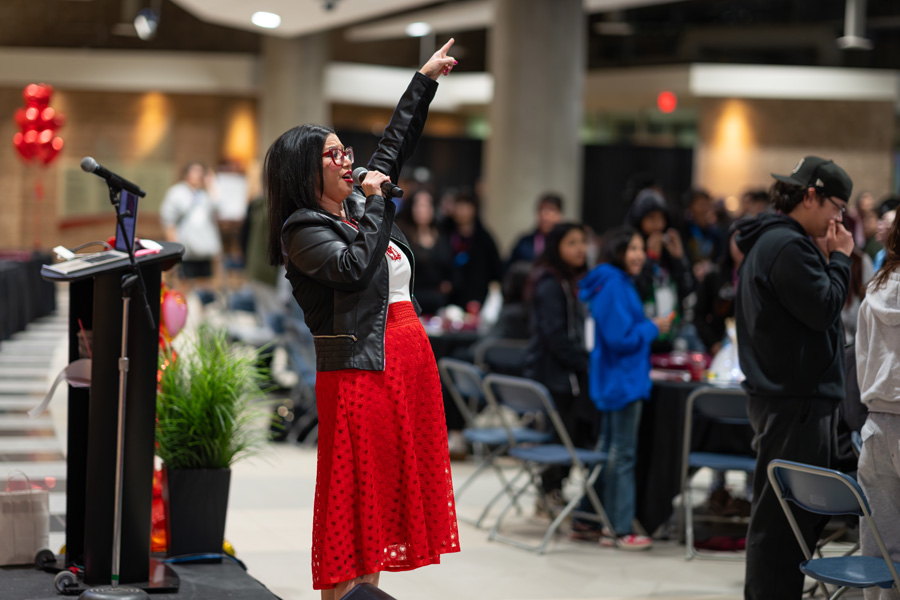 A woman in a black jacket and red skirt holds her hand up while speaking to a room of young people